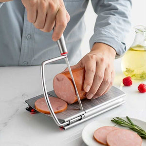 Stainless steel cheese slicer with a silver cutting wire and measurement grid, slicing pink ham with red anti-slip feet