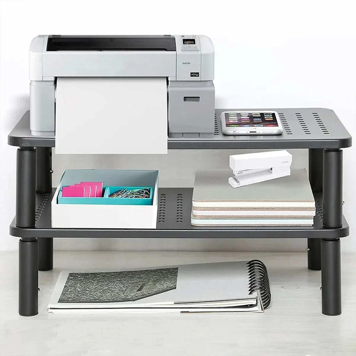 Black iron Adjustable Monitor & Laptop Riser with two perforated shelves and sturdy cylindrical legs, holding office supplies and devices.