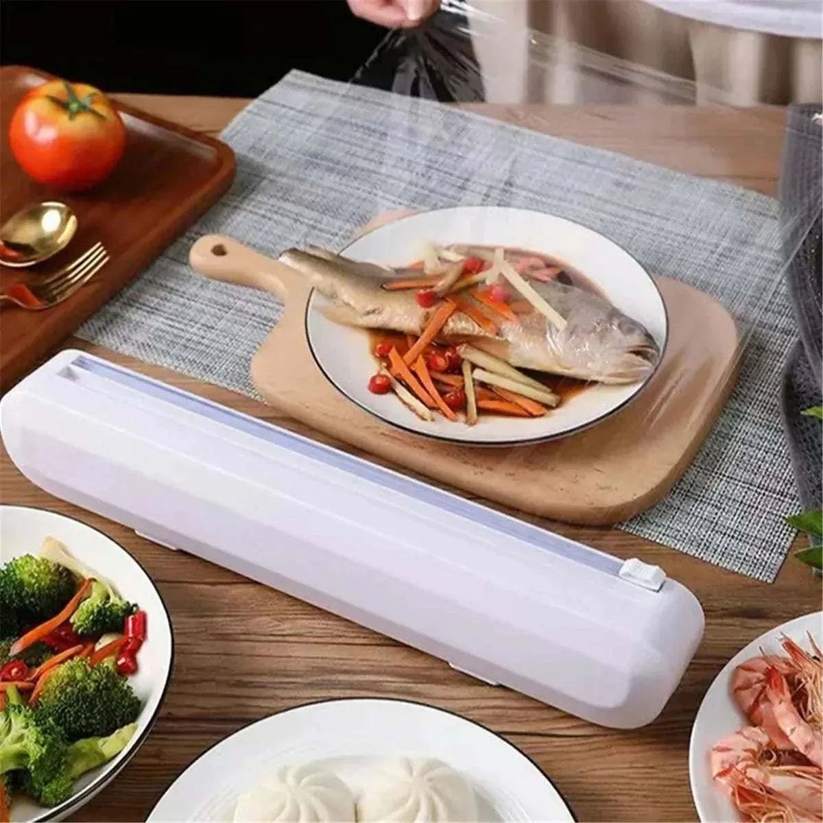 White plastic Cling Film Cutter Box with a suction cup base on a wooden table covering a plate of fish and vegetables with cling film