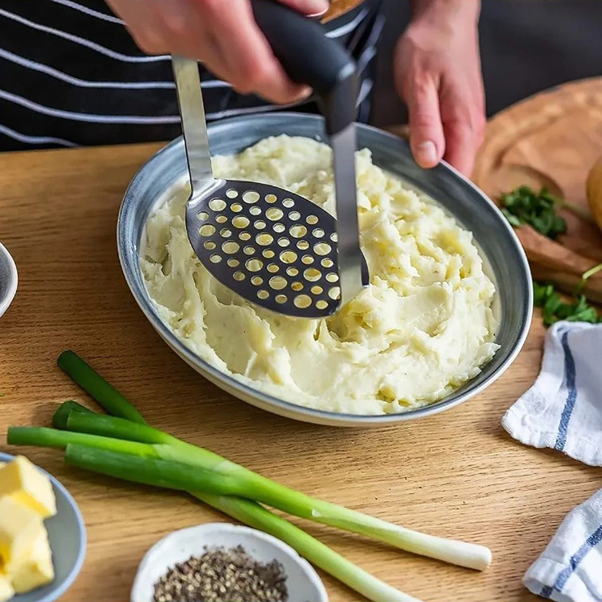 Stainless Steel Potato Masher with a perforated metal plate and black wide grip, pressing creamy mashed potatoes