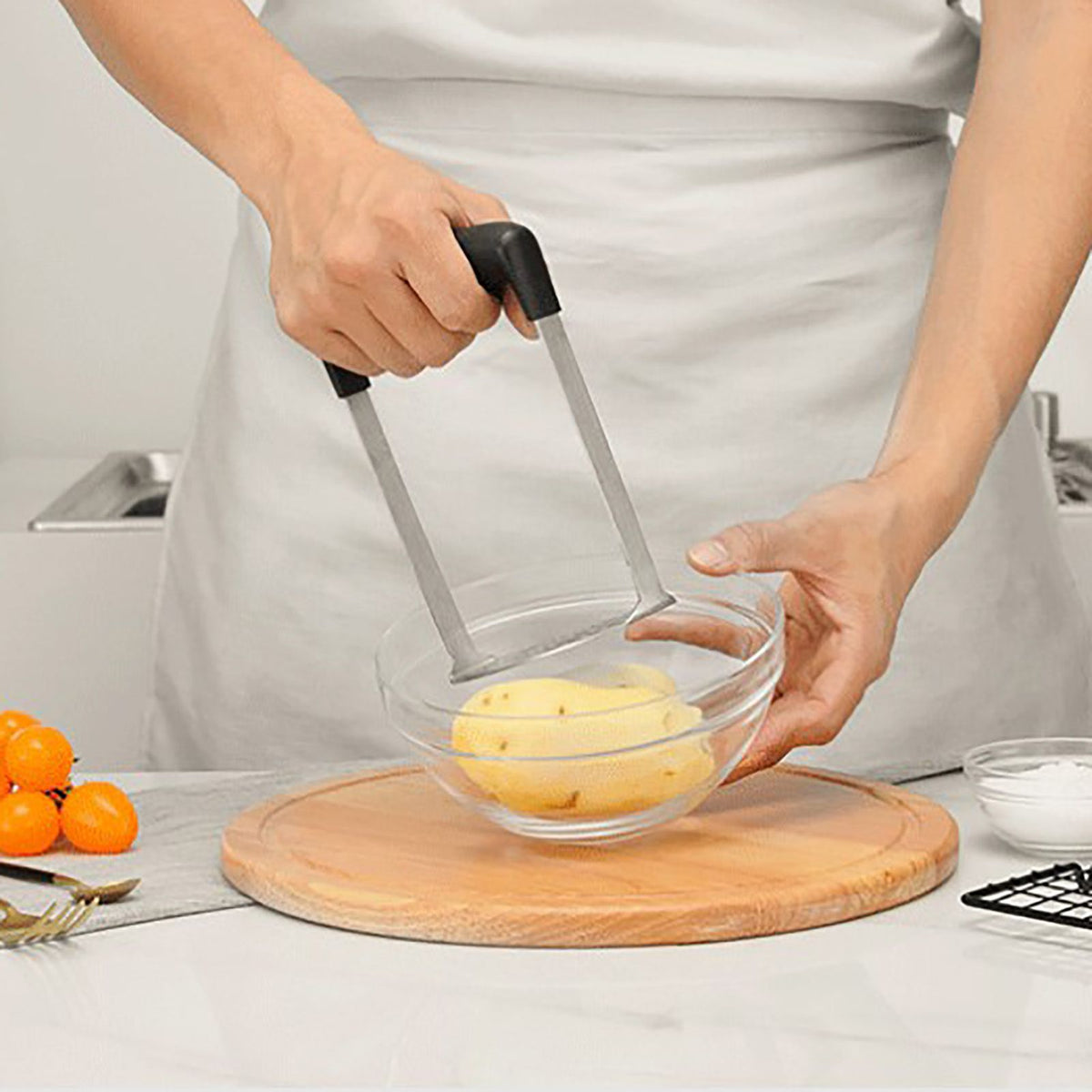 Stainless Steel Potato Masher with a wide metal mashing plate and black grip handles pressing a cooked potato in a glass bowl